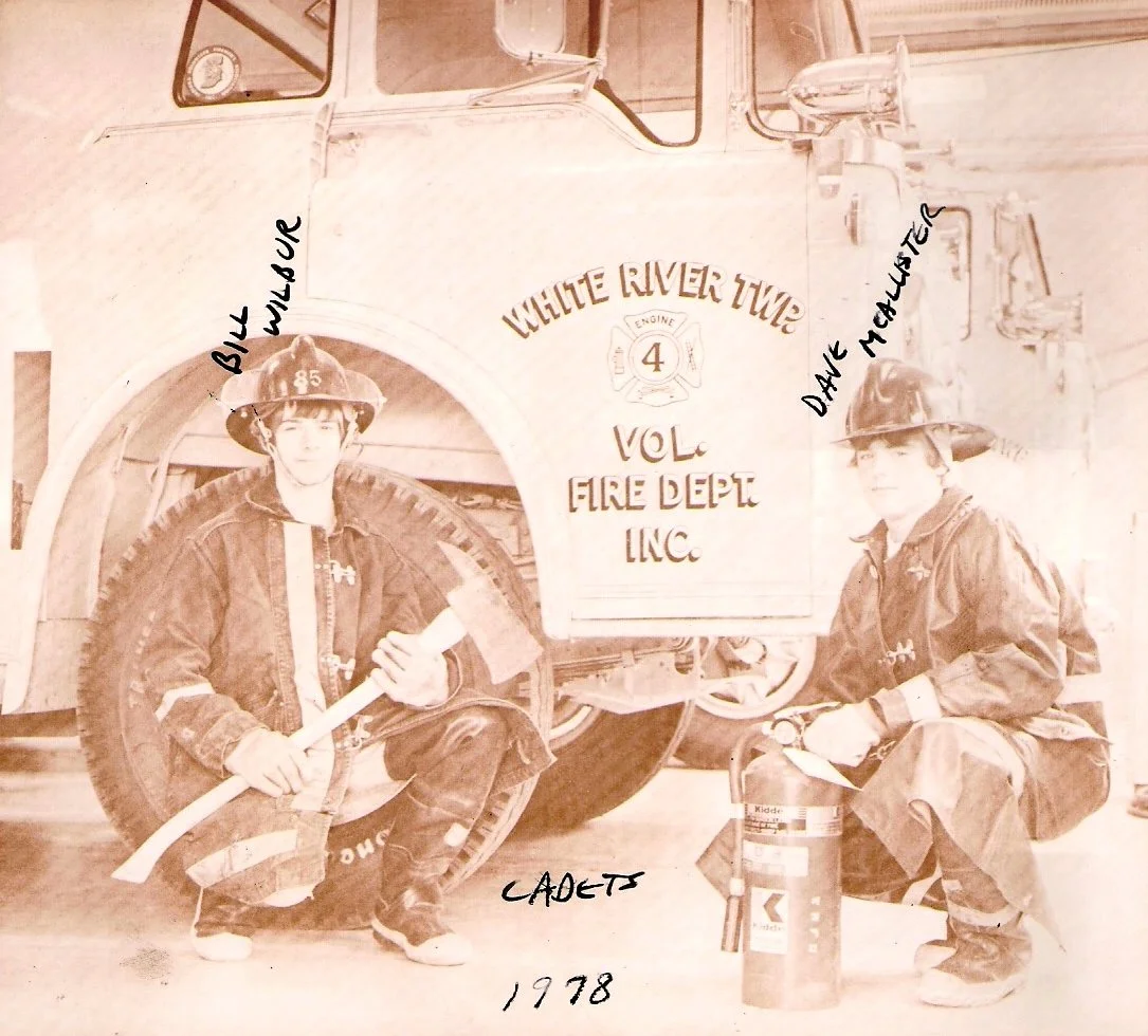 A sepia-toned vintage photograph shows two young firefighters in uniform, kneeling next to a fire truck. The fire truck has the words 'White River Tour Vol. Fire Dept. Inc.' painted on it. One firefighter is holding a rolled-up fire hose, and the oth