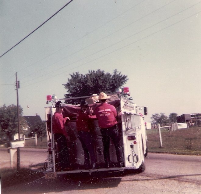 Three firefighters in red shirts and hats standing on a fire truck on a rural street, with houses and trees in the background.
