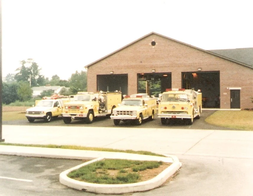 Five yellow emergency vehicles parked outside a fire station with three garage doors.