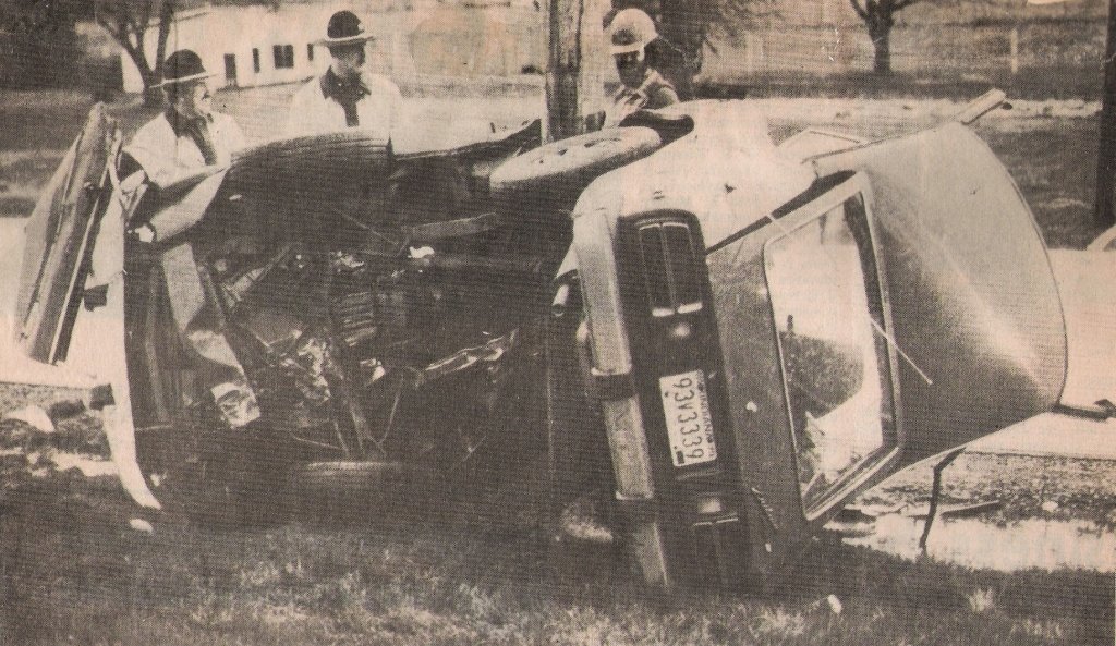 A black and white photo of a car accident with a flipped-over vehicle and three people standing nearby, possibly police officers or investigators.
