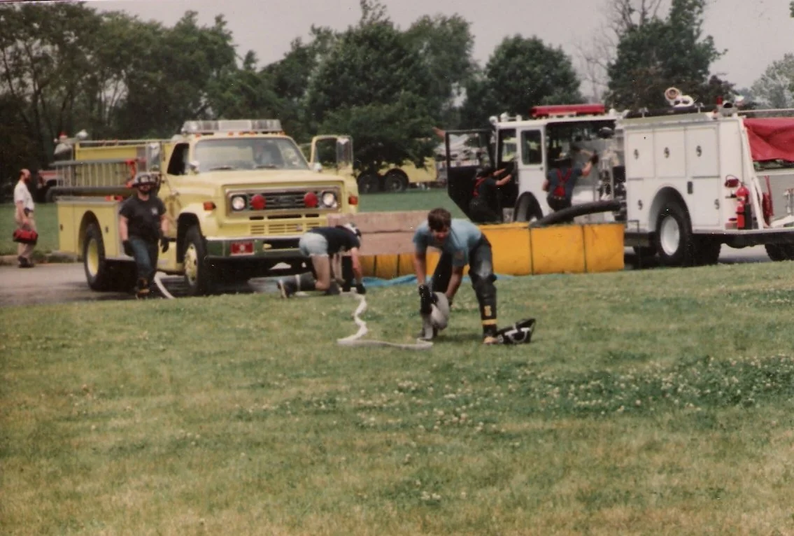 Firefighters training with fire trucks and hoses on a grassy field with trees in the background.