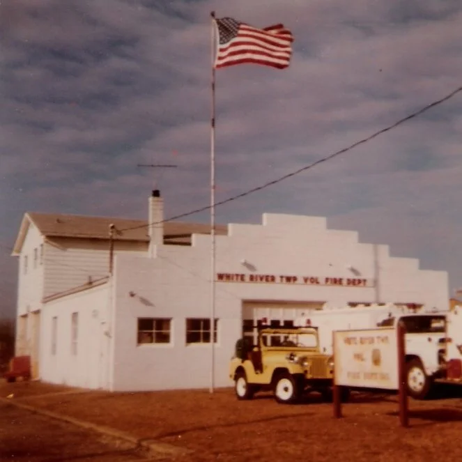 American flag flying in front of a white fire department building with a sign that reads 'White River TWP Vol Fire Dept.' Two fire trucks are parked outside in a grassy area.
