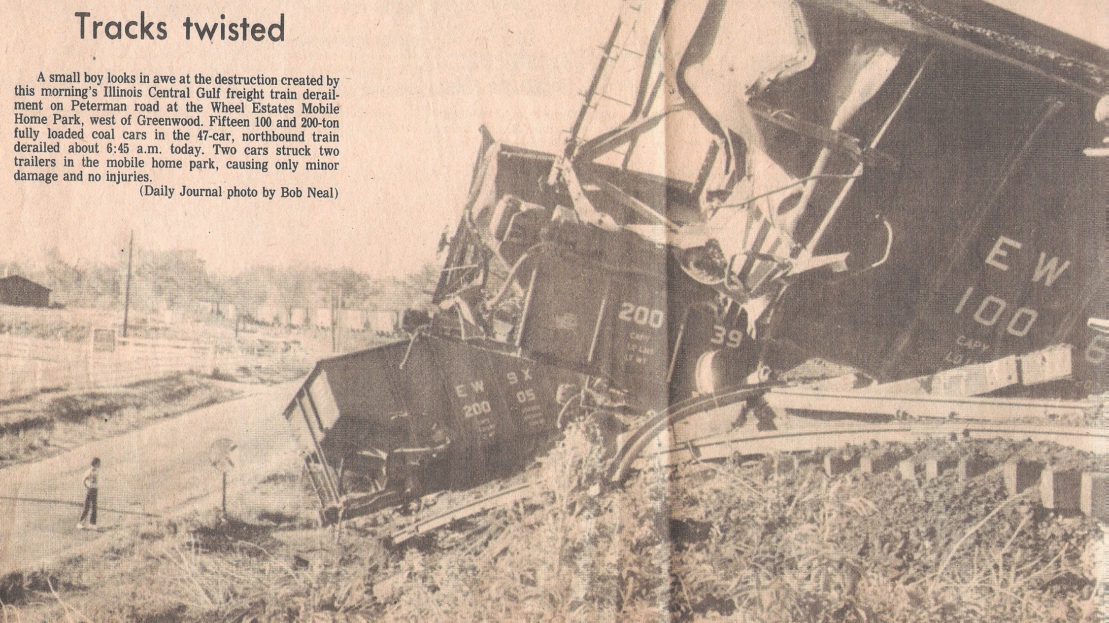 A train derailed on tracks with debris scattered around. An individual stands nearby observing the scene, with utility poles and buildings in the background.