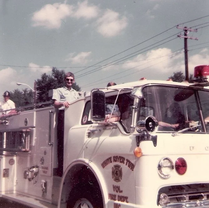 Vintage fire truck with a person standing in the driver’s seat and others on top, during daytime with partly cloudy sky and power lines in the background.
