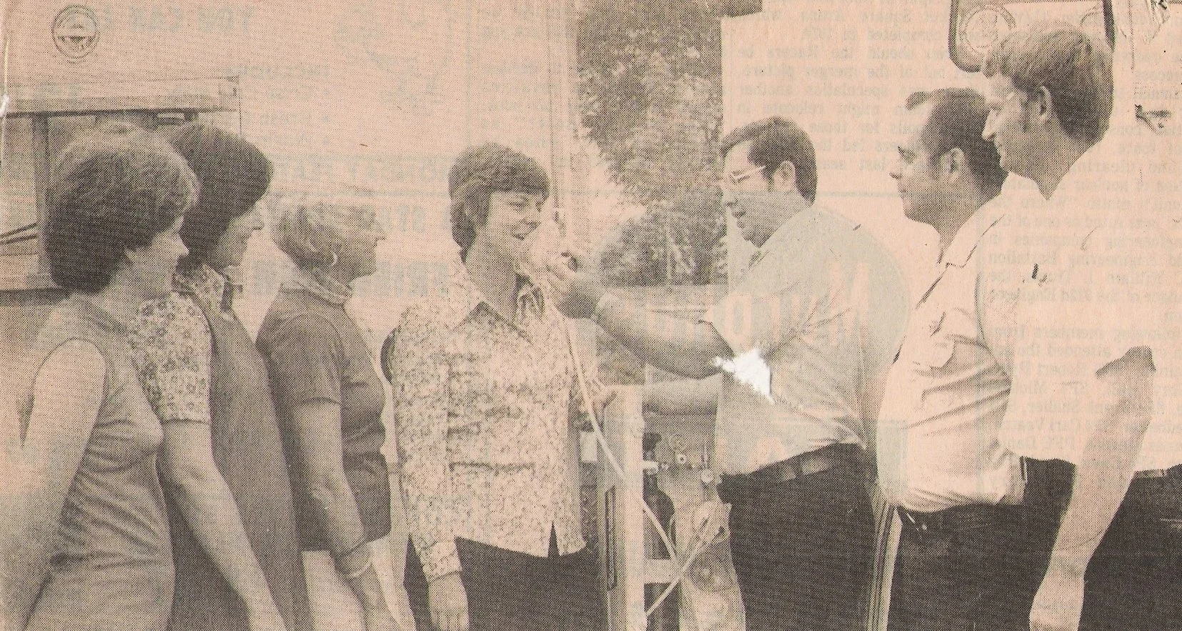 A historical black-and-white photograph showing a group of seven people, five women and two men, standing outdoors. The group appears to be engaged in a medical or health-related activity, with one woman receiving a checkup or health measurement from
