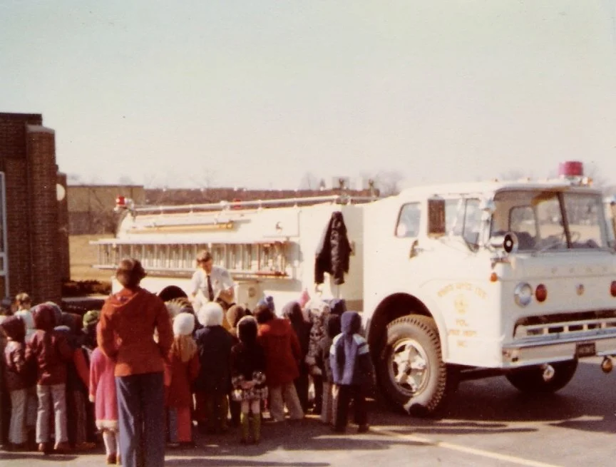 A vintage fire truck stopped on a street with a crowd of children and adults gathered around it, during what appears to be a community event outdoors.