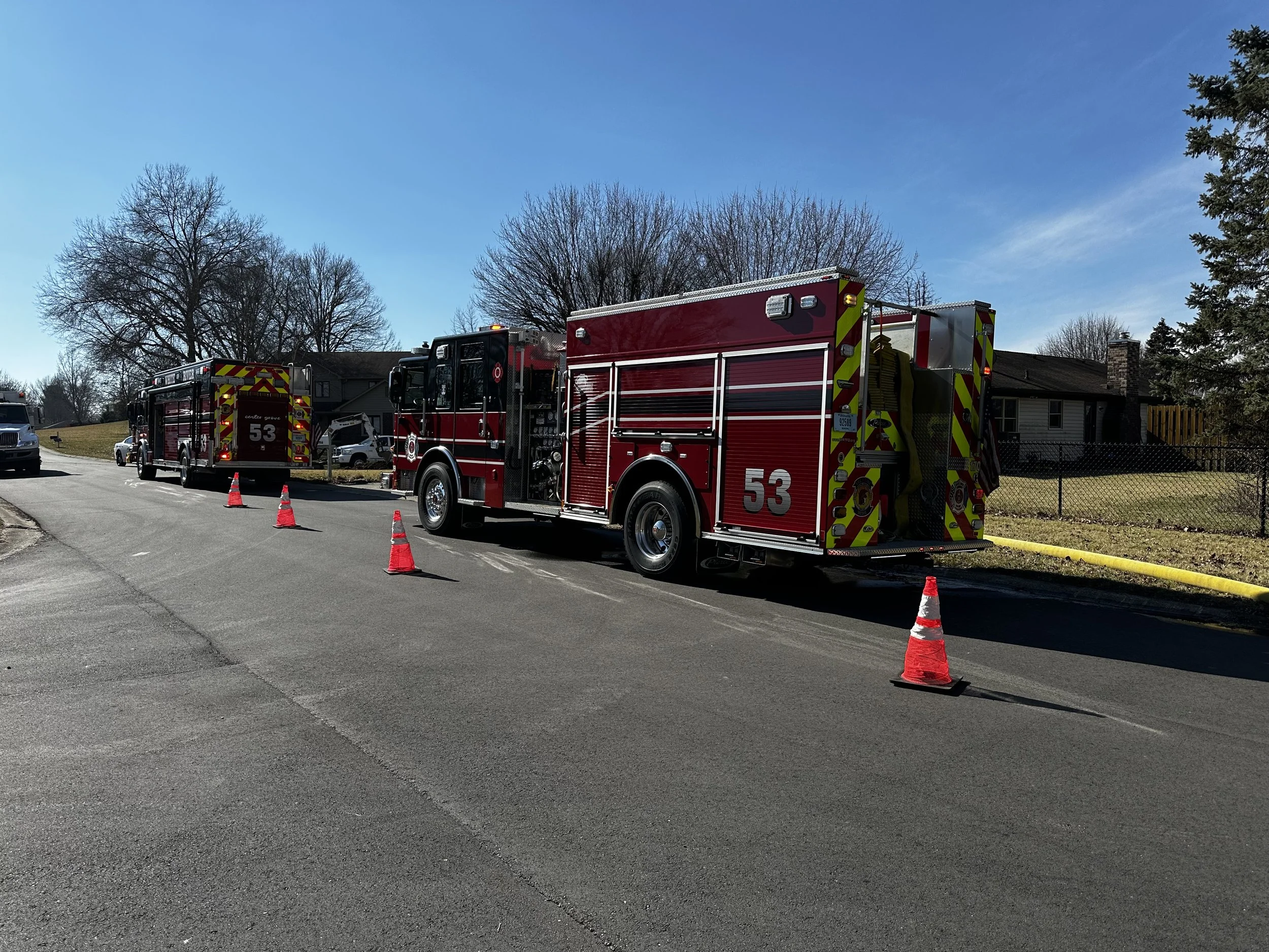 Two red fire trucks parked on the side of a street, with traffic cones placed around them, and a house with trees in the background.