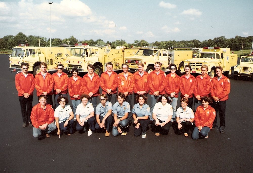 Group of young emergency responders, including firefighters and paramedics, standing outdoors in front of yellow fire trucks and emergency vehicles, wearing orange and light blue uniforms.