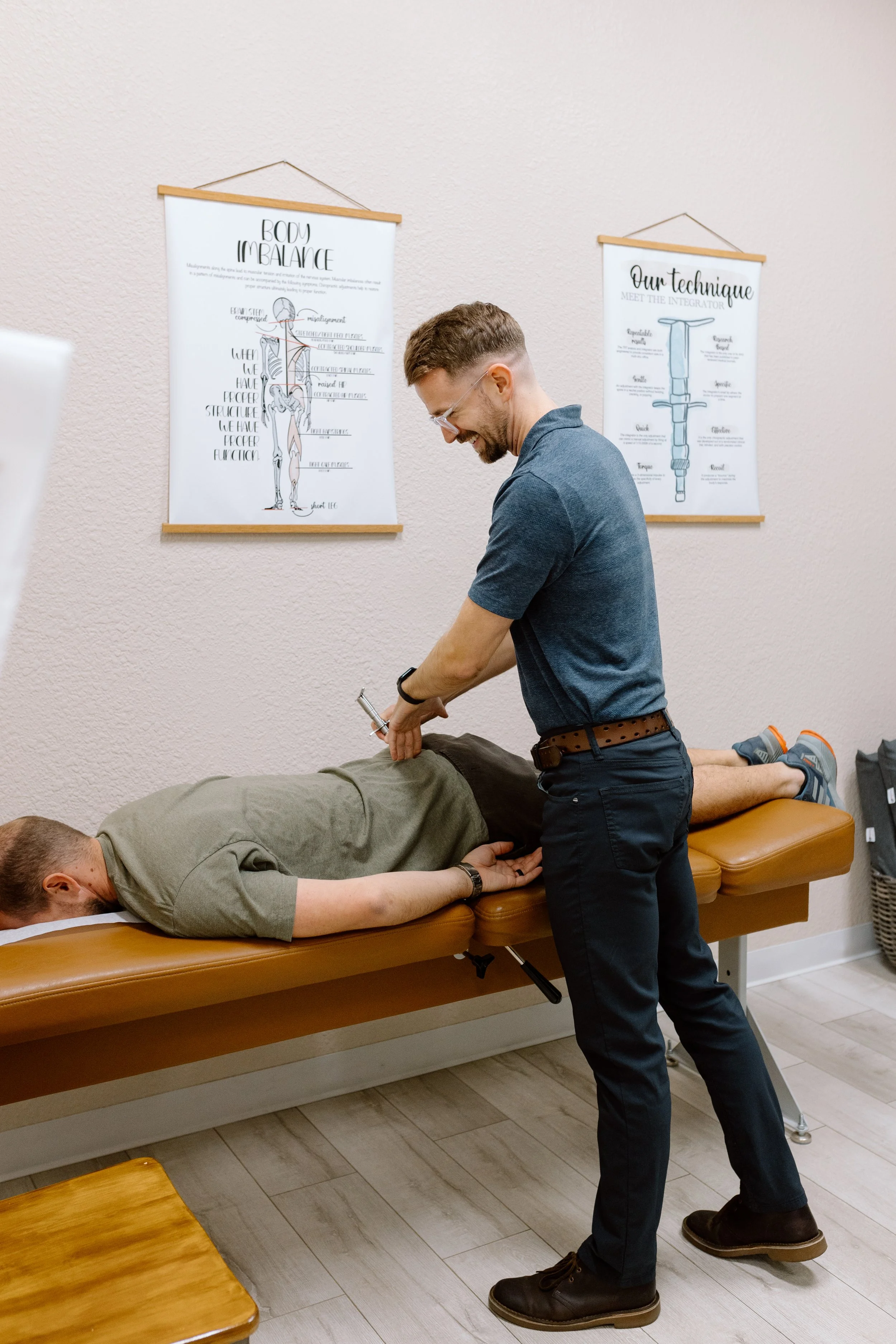 A chiropractor adjusting a patient's lower back in an examination room with chiropractic posters on the wall.