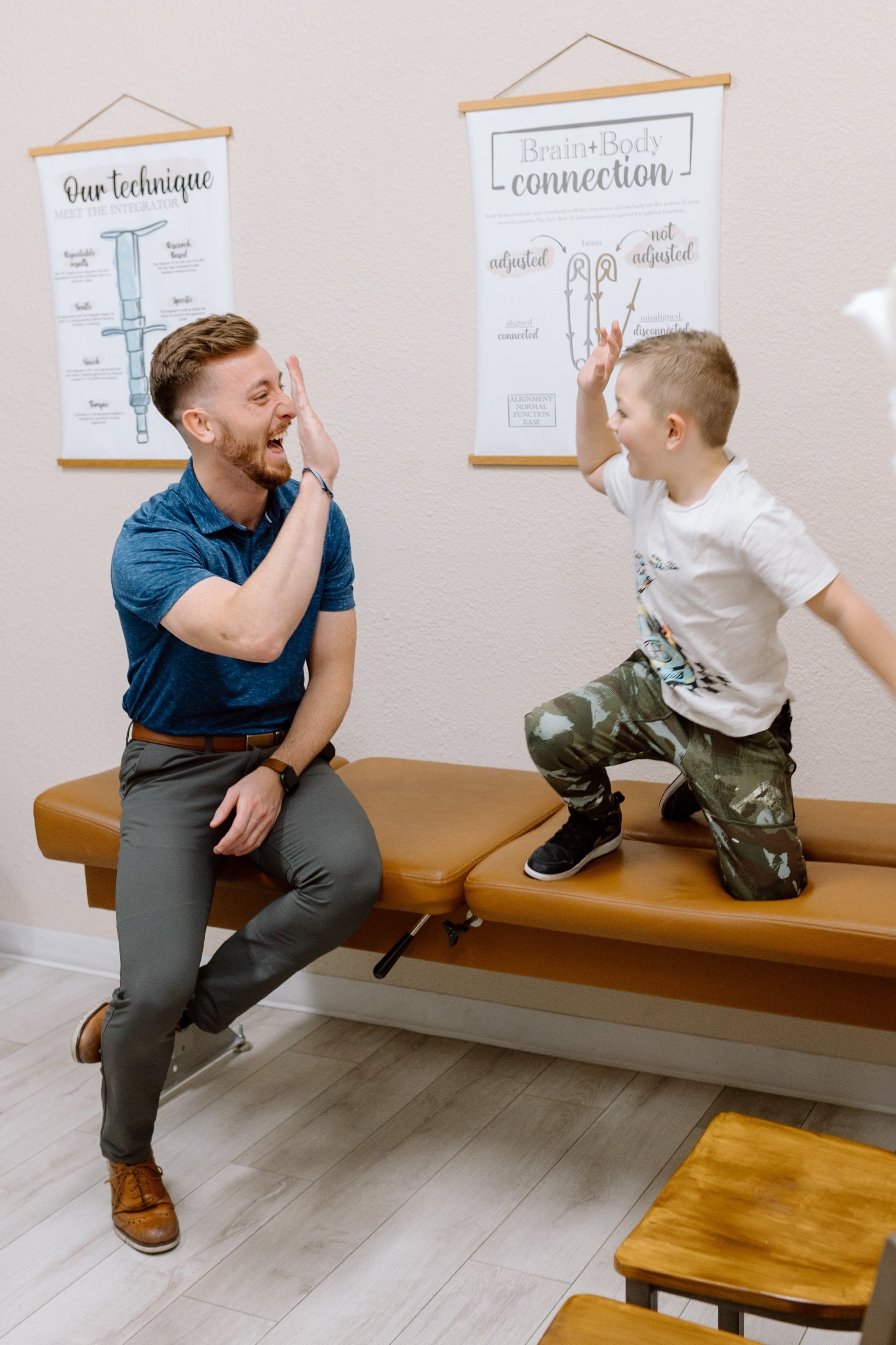A man and a young boy are giving each other high fives in a chiropractic therapy setting, with educational posters on the wall behind them.