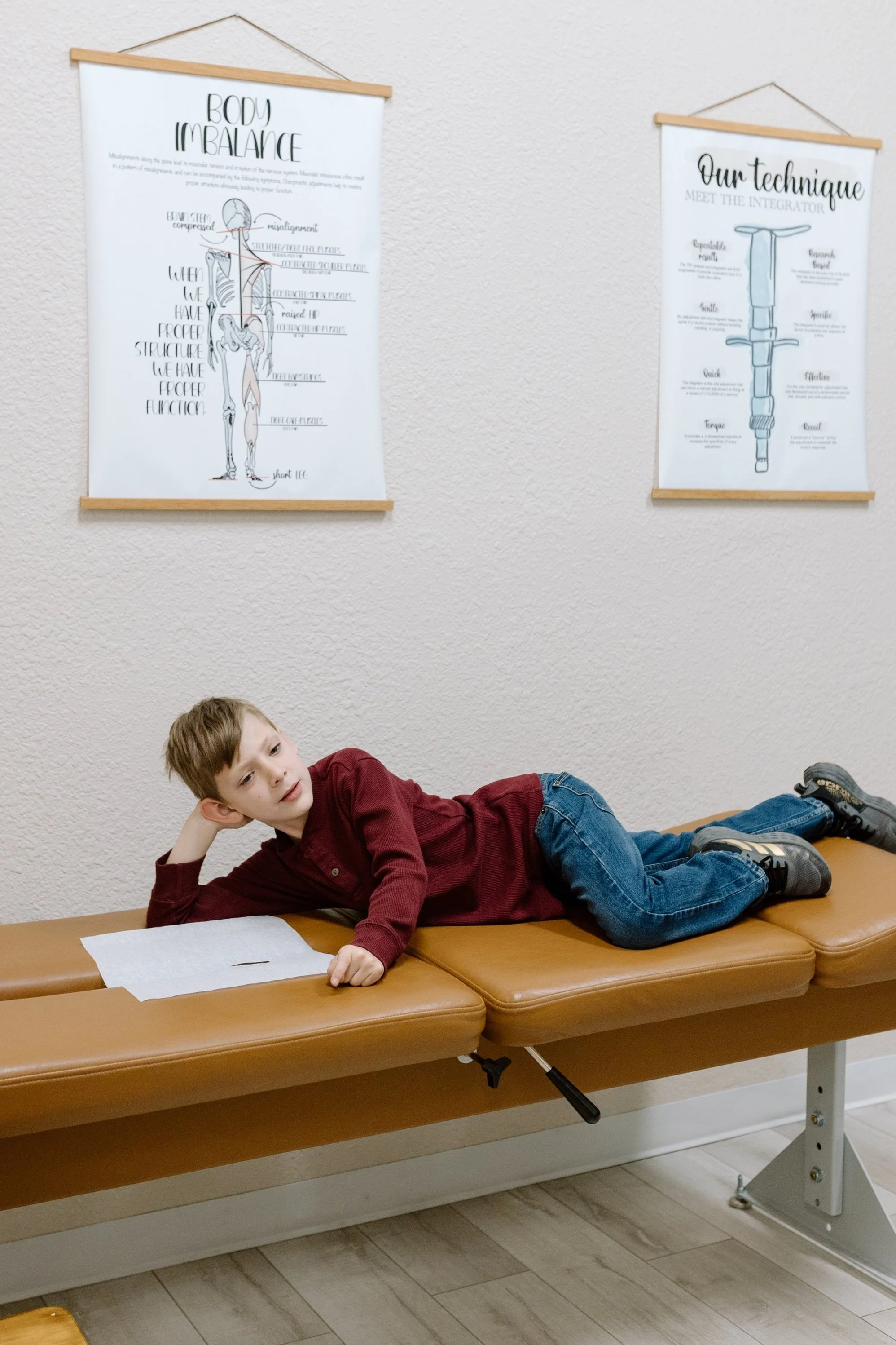 A young boy lying on an examination table in a chiropractic office, with educational posters about body balance and technique hanging on the wall behind him.