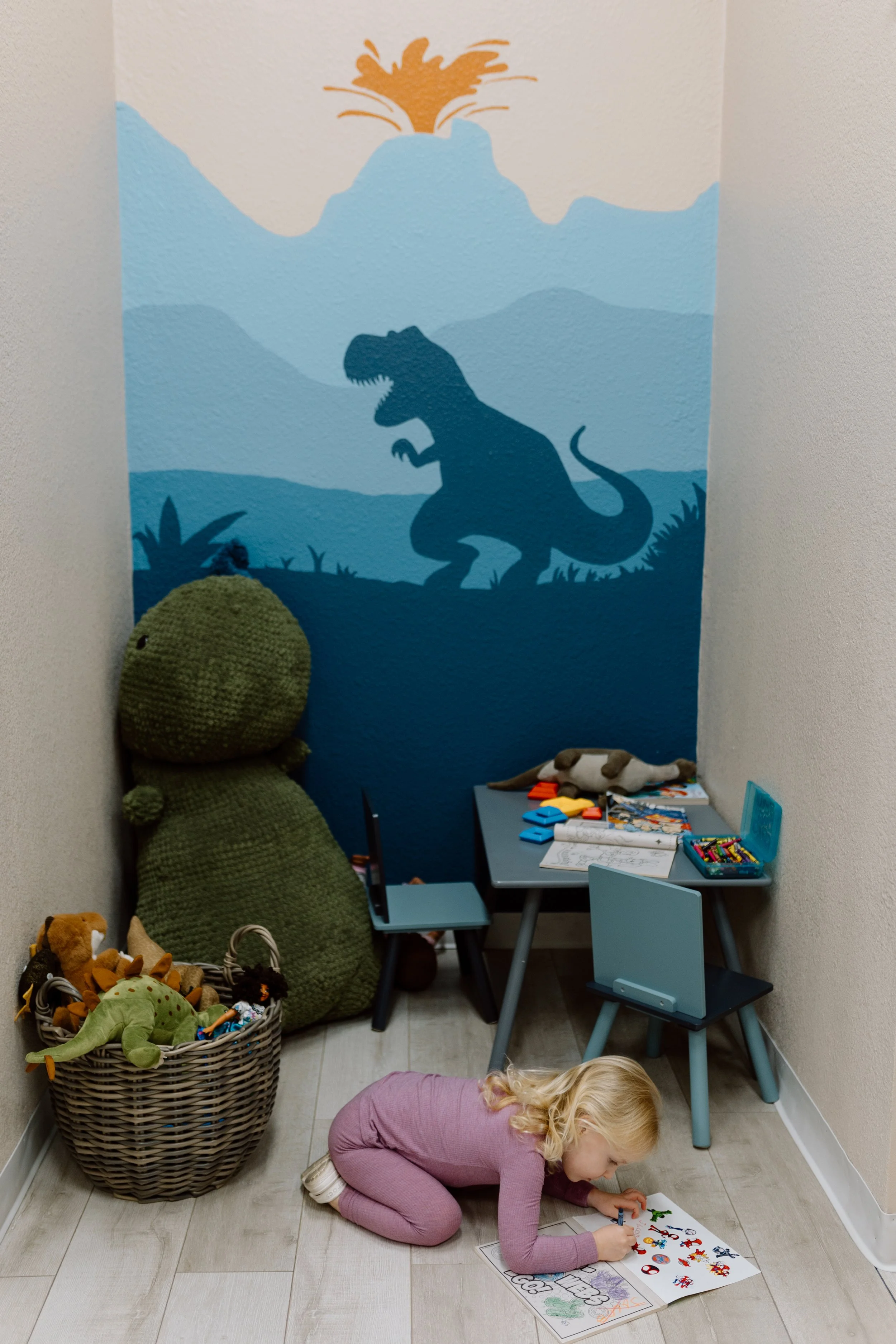 A girl with blonde hair in pink clothes kneeling on the floor coloring a dinosaur poster in a playroom. The room has a blue dinosaur mural on the wall, a large stuffed dinosaur, and a basket of stuffed animals.
