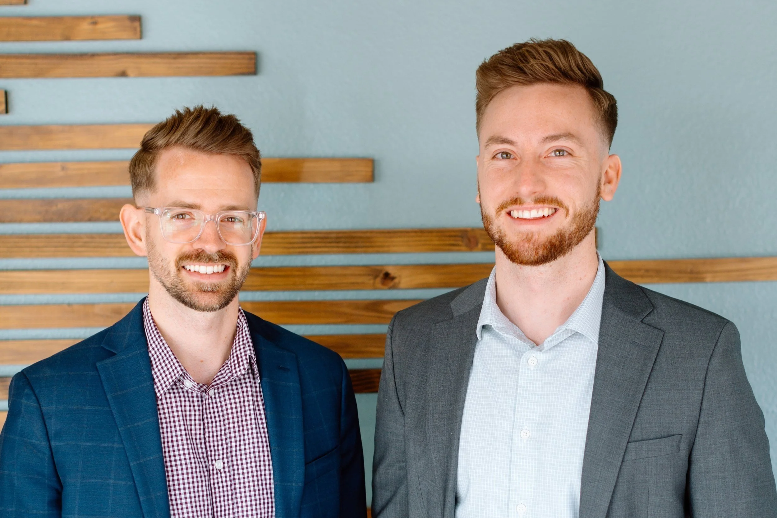 Two young men in business suits standing indoors, smiling at the camera, with a modern wooden wall in the background.