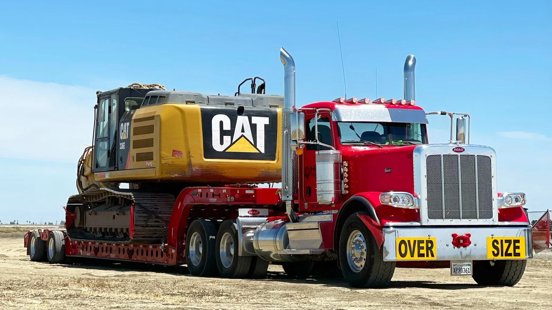 A large semi-truck H&S Heavy Haul with a red cab and a yellow and black Caterpillar excavator on a flatbed trailer. The trailer has a sign indicating the truck is over size.