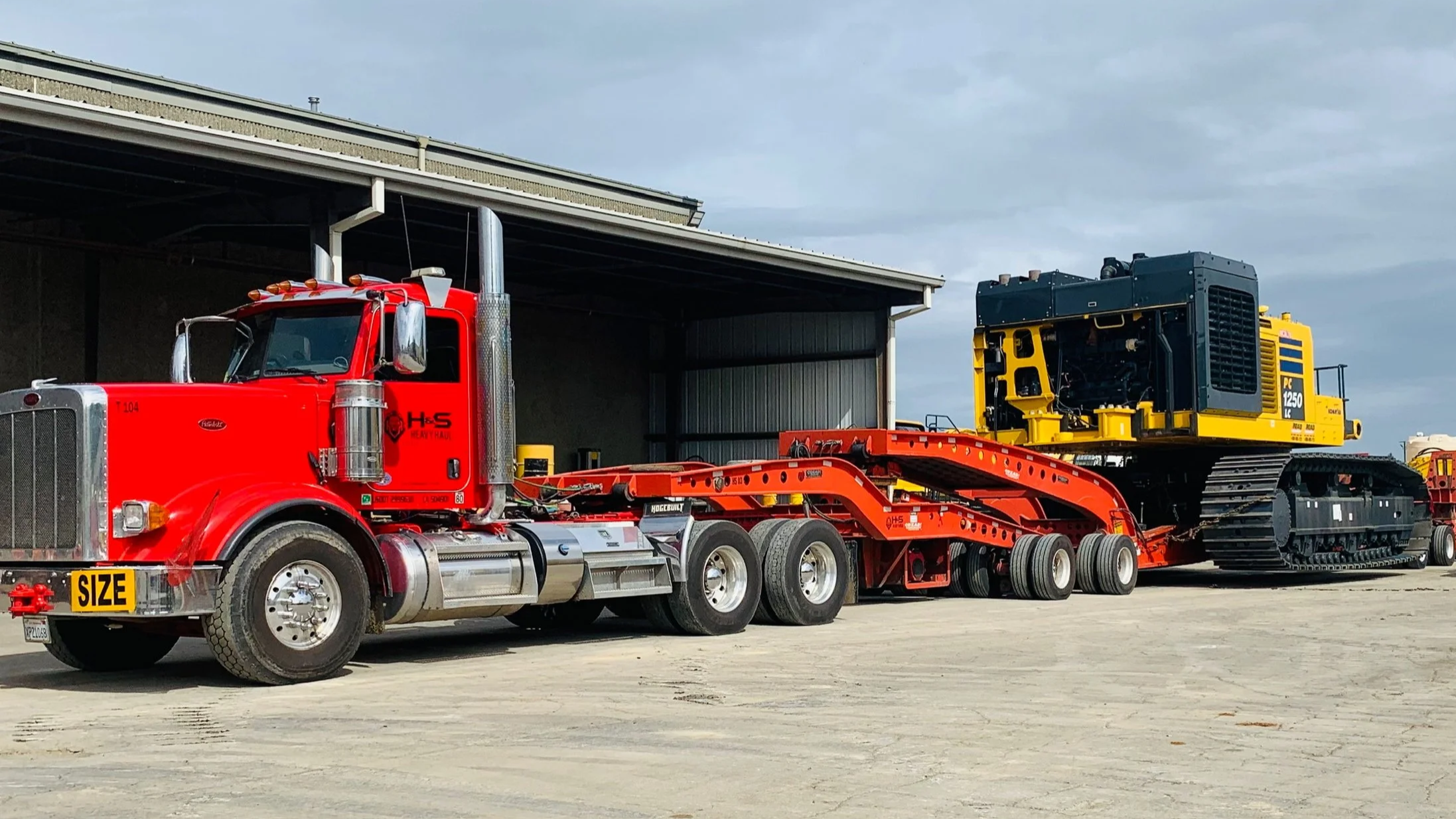 H&S Heavy Haul, The image shows a large red heavy-duty truck with a flatbed trailer carrying a yellow and black tracked construction equipment. The truck is parked outside a metal building with a partially open garage door.
