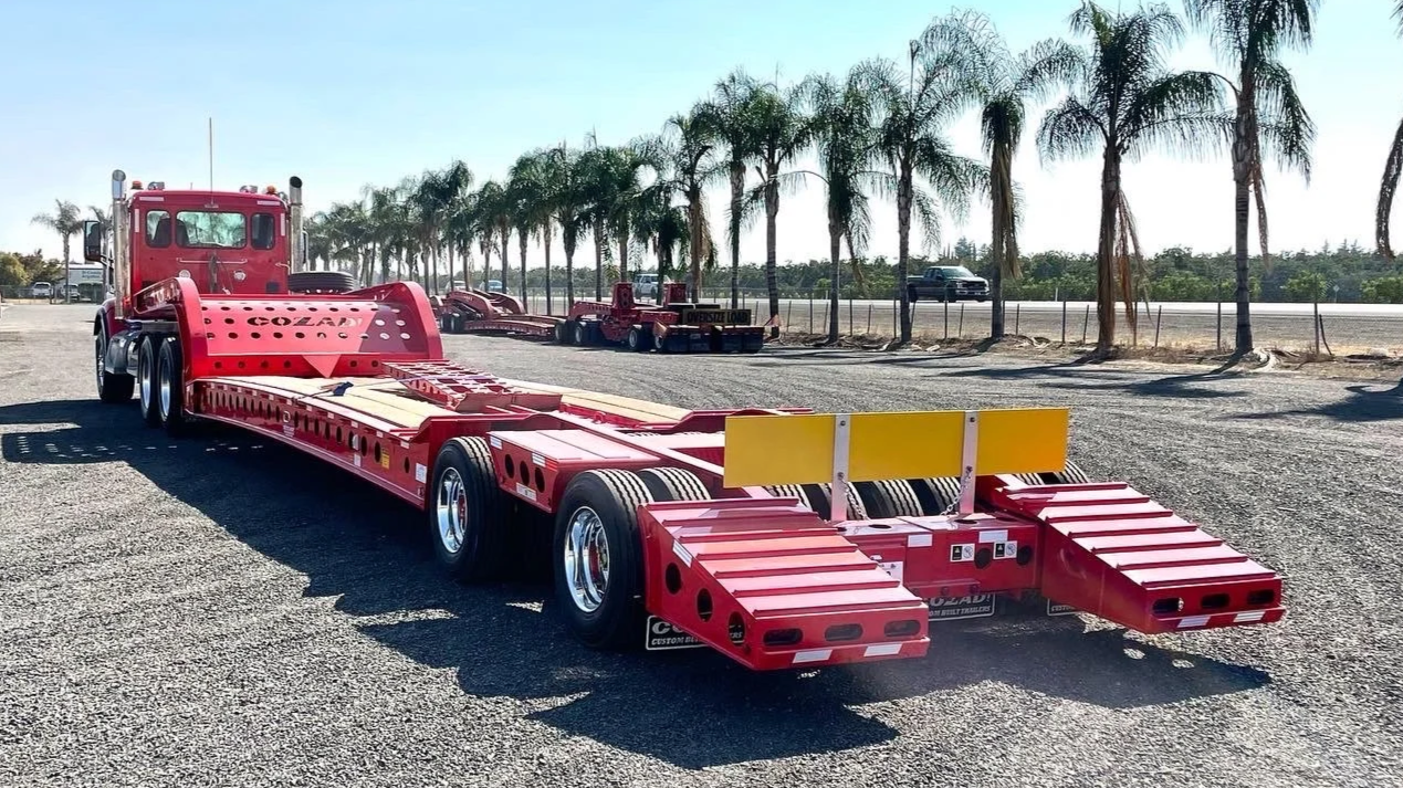 H&S Heavy Haul, Large red flatbed truck trailer parked on gravel with palm trees and a highway in the background.