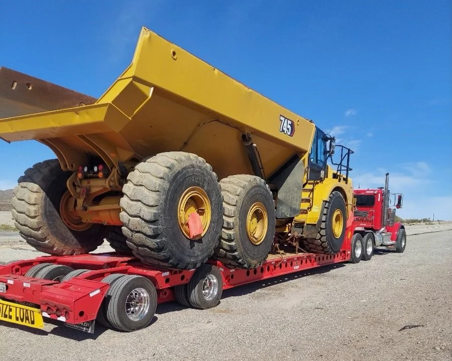 A yellow construction dump truck loaded on a red flatbed trailer from H&S Heavy Haul being transported on a highway with a clear blue sky in the background.