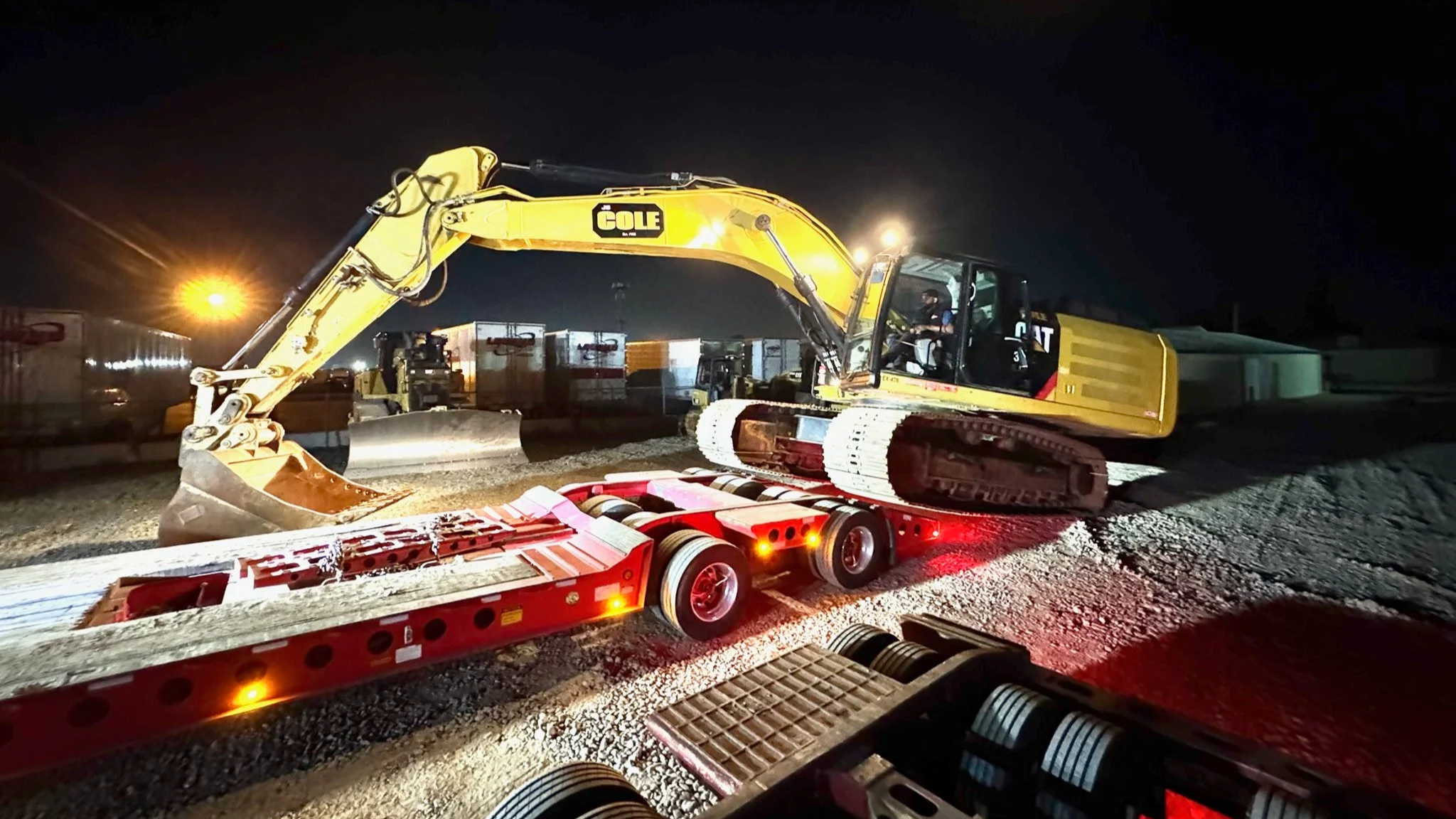 H&S Heavy Haul Nighttime construction scene with a yellow excavator on a red trailer, working on a gravel surface. Bright lights illuminate the area, with containers and buildings in the background.