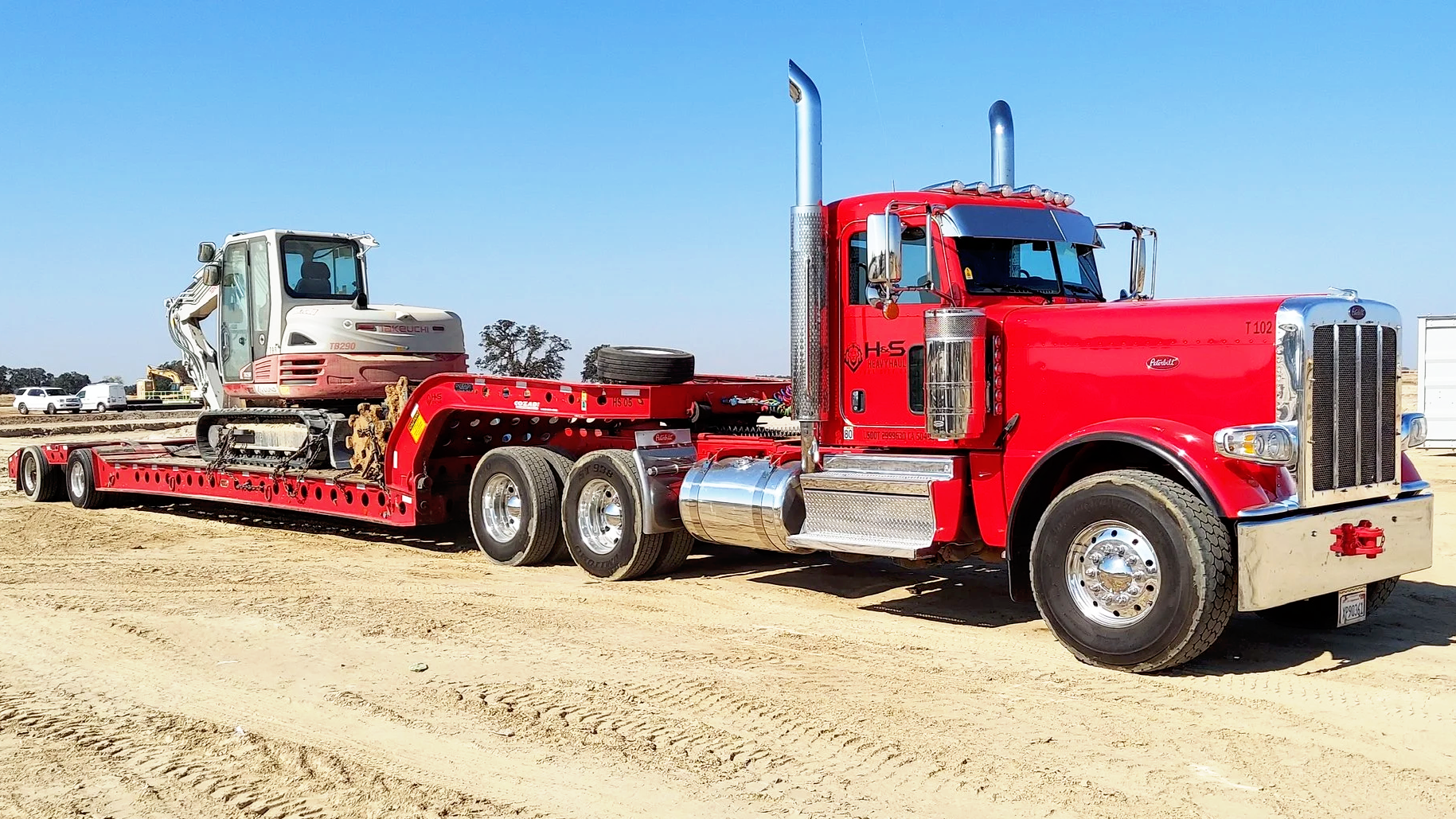 Red flatbed truck from H&S Heavy Haul carrying a white excavator on a dirt lot with vehicles and trees in the background.