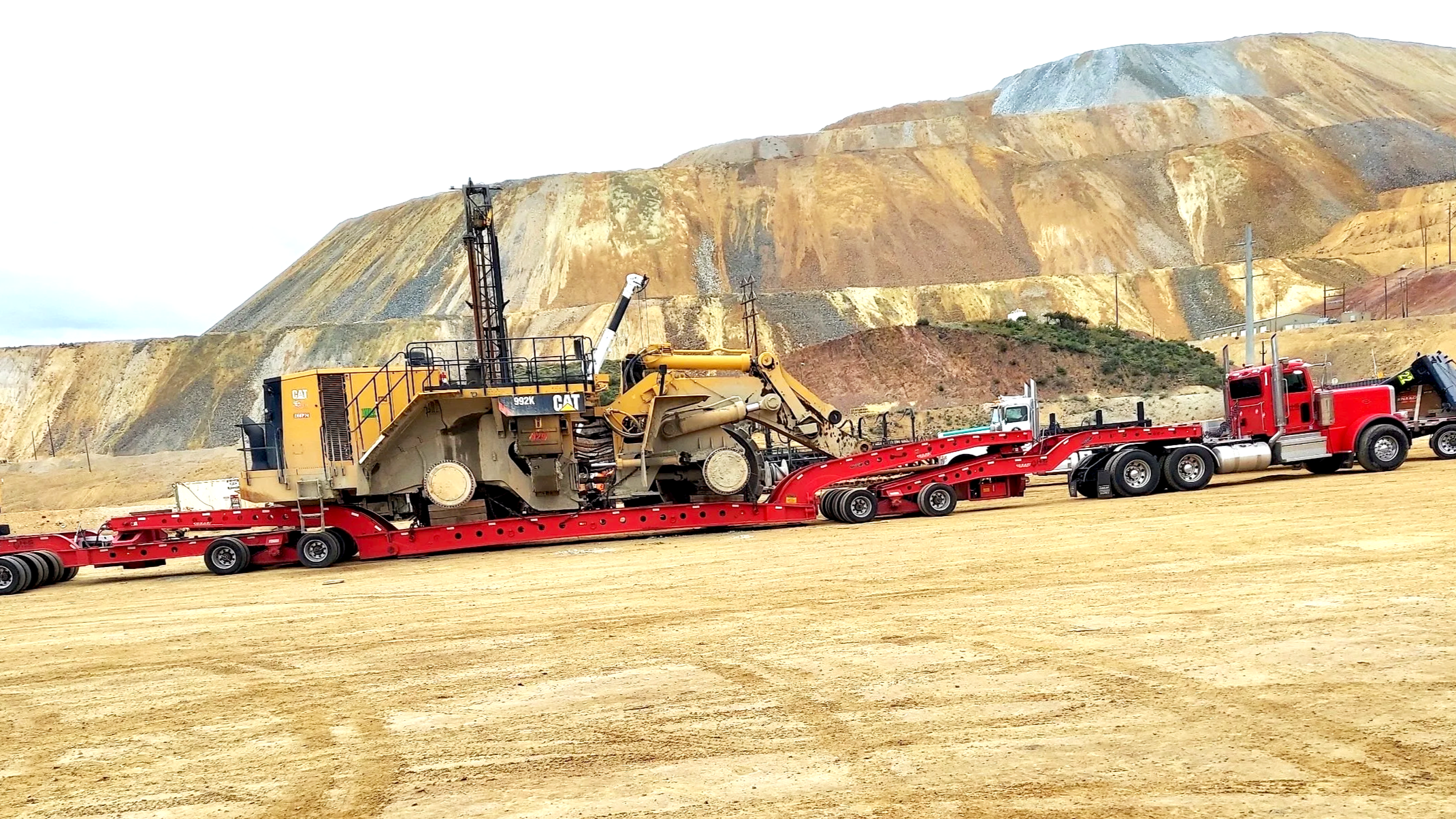 A large yellow Caterpillar 992K wheel loader on a flat red transport trailer from H&S Heavy Haul in an open land with a colorful mined mountain in the background.