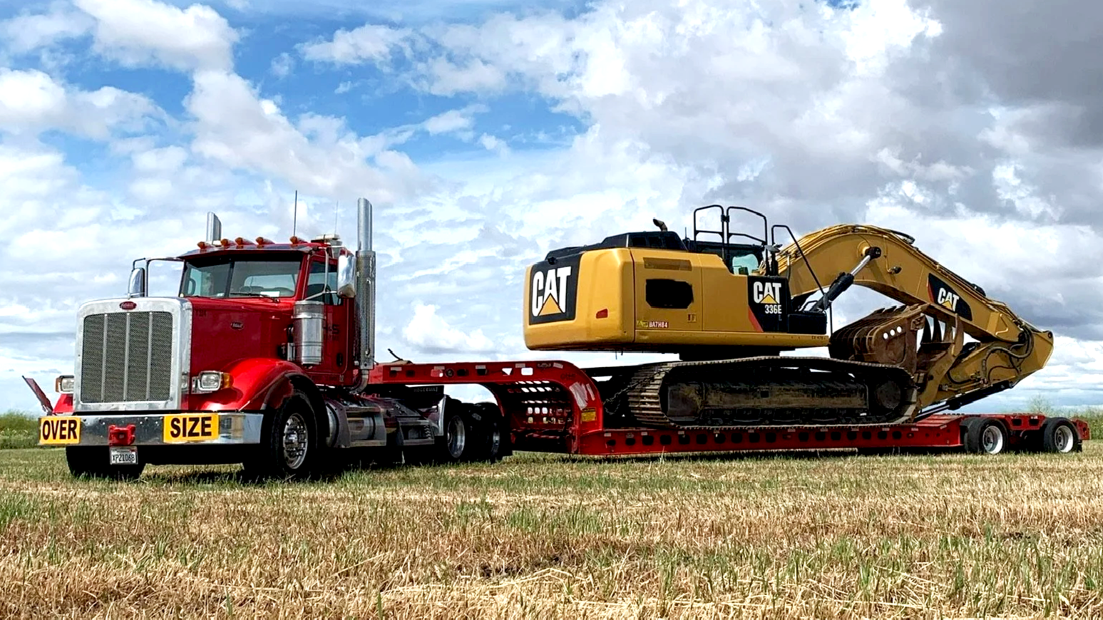 A red semi-truck from H&S Heavy Haul carrying a yellow CAT excavator on a flatbed trailer in an open field with cloudy sky.