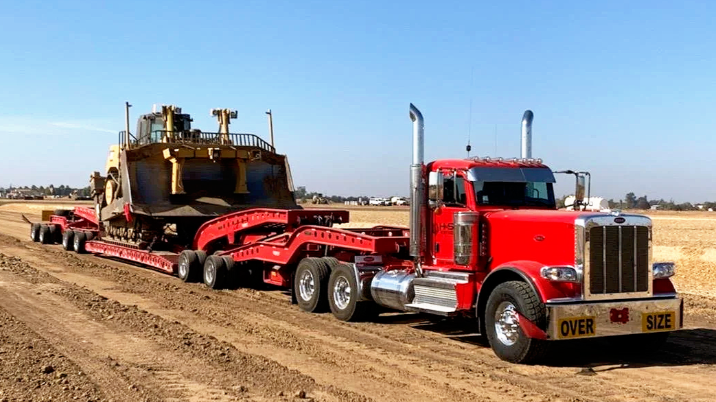 A red semi-truck H&S Heavy Haul transporting a large, yellow bulldozer on a flatbed trailer across a dirt road in a rural area with open fields and a clear blue sky.