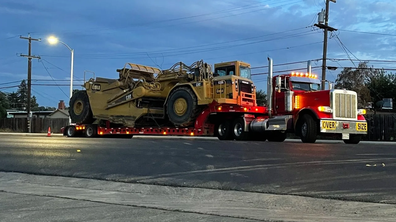A large red semi-truck from H&S Heavy Haul with an oversized load sign is transporting a yellow bulldozer on a flatbed trailer during dusk, with overcast sky and power lines in the background.