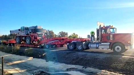 A large red flatbed tow truck from H&S Heavy Haul transporting a dark gray vehicle on a highway with clear skies and trees in the background.