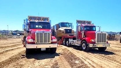 Two large red trucks from H&S Heavy Haul with signs that read "Oversize Load" on top, parked on a dirt road in a rural area during daytime.