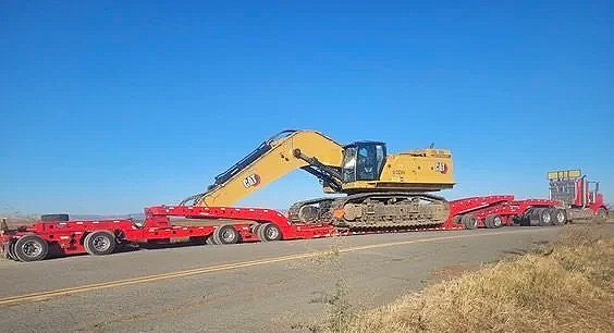 A yellow excavator being transported on a flatbed semi-truck from H&S Heavy Haul along a highway with clear blue sky and dry grass.