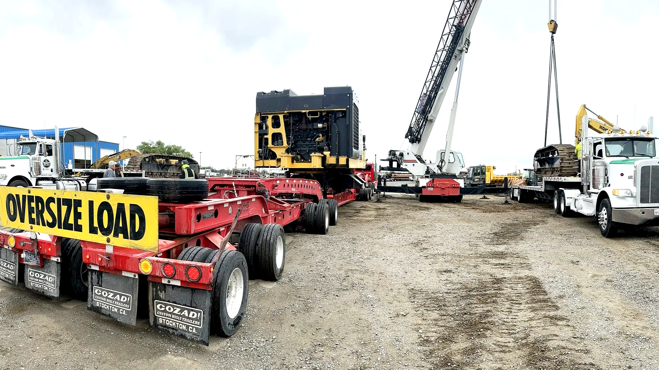 Construction site with trucks from H&S Heavy Haul, cranes, and heavy machinery, including a large black and yellow machine being lifted by a crane, and a yellow sign reading 'OVERSIZE LOAD' in the foreground.