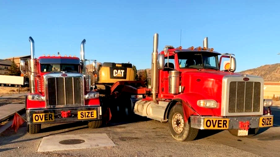 Two red semi-trucks from H&S Heavy Haul with 'Over Size' signs on front bumpers parked side by side on a paved lot against a clear blue sky.