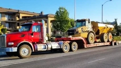 A red semi-truck from H&S Heavy Haul  with a flatbed trailer carrying a large yellow dump truck parked on a street.