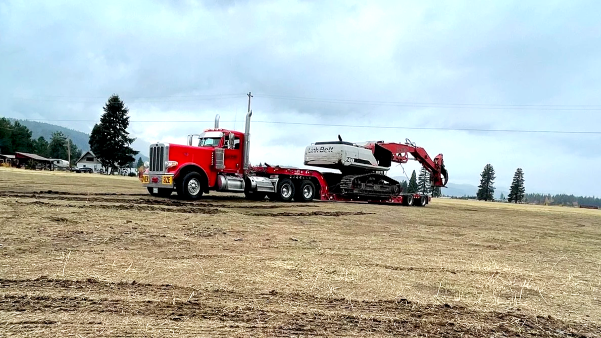 A large red truck from H&S Heavy Haul pulling a tracked excavator on a flat, open field with a rural background of trees and houses under a cloudy sky.