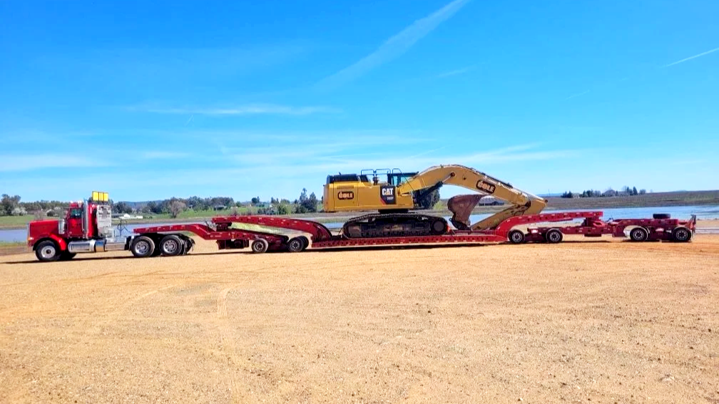 A flatbed truck from H&S Heavy Haul transporting a large yellow excavator near a body of water on a sunny day.