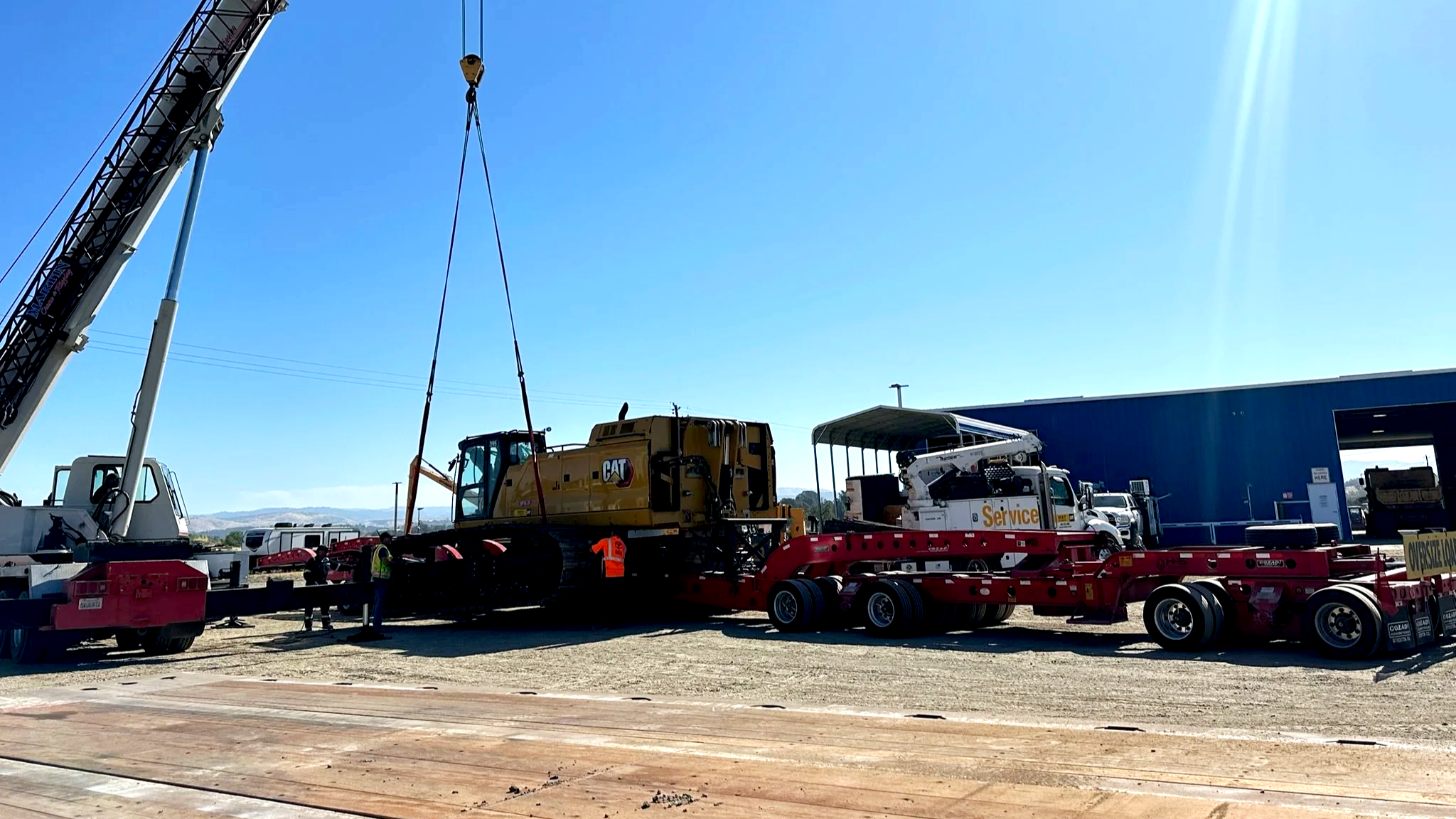 Construction site with a large crane lifting a yellow Caterpillar excavator onto a red transport trailer from H&S Heavy Haul, with workers present and a blue warehouse in the background.