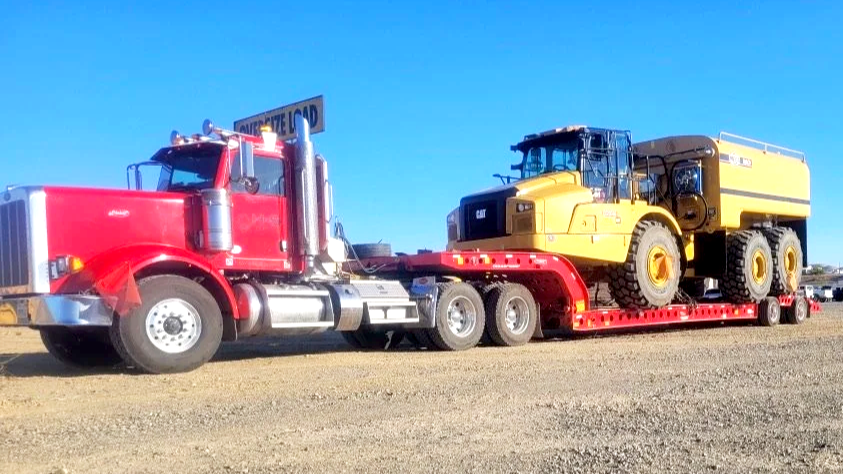 A red semi-truck from H&S Heavy Haul transporting a large yellow Caterpillar road roller on a flatbed trailer.