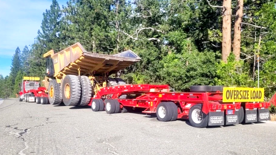 A loaded semi-truck from H&S Heavy Haul transporting a large, yellow mining dump truck on a flatbed trailer on a rural road with trees in the background.