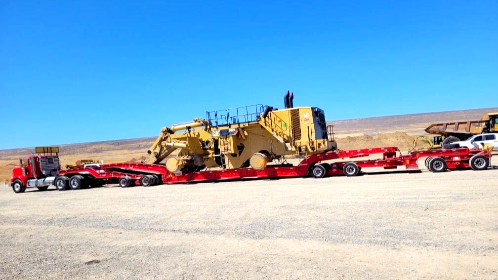 A large red flatbed trailer from H&S Heavy Haul transporting a yellow Caterpillar excavator in an open desert area under a clear blue sky.