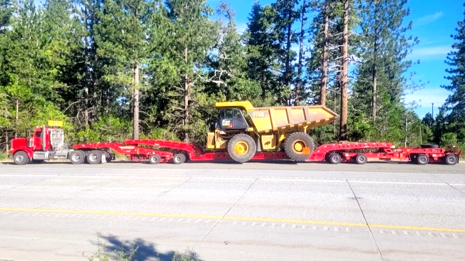 A large yellow dump truck is loaded onto a red flatbed trailer, which is attached to a red semi-truck from H&S Heavy Haul, parked on the side of a road with trees and a blue sky in the background.