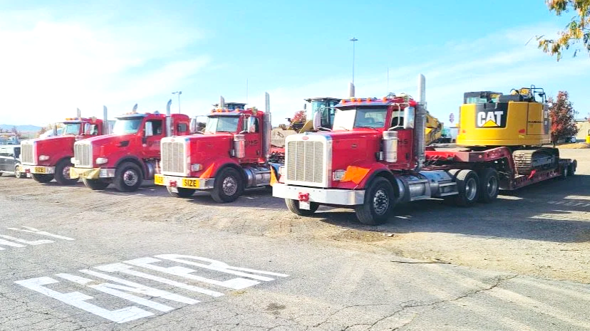 A row of four red semi-trucks from H&S Heavy Haul parked side by side, with a yellow Caterpillar bulldozer on a trailer attached to the last truck, in a parking lot. 