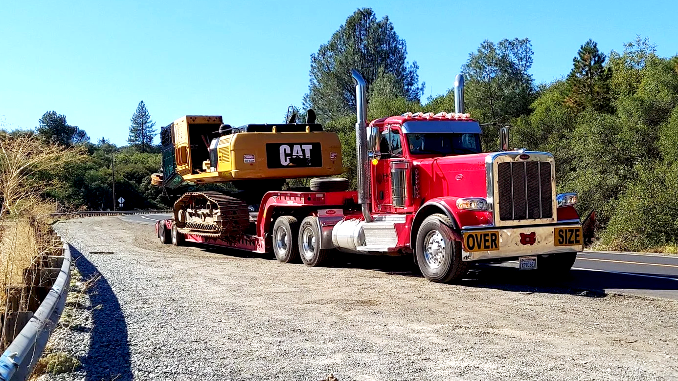 A red semi-truck from H&S Heavy Haul transporting a large yellow CAT excavator on a flatbed trailer on a rural road with trees and a clear blue sky.