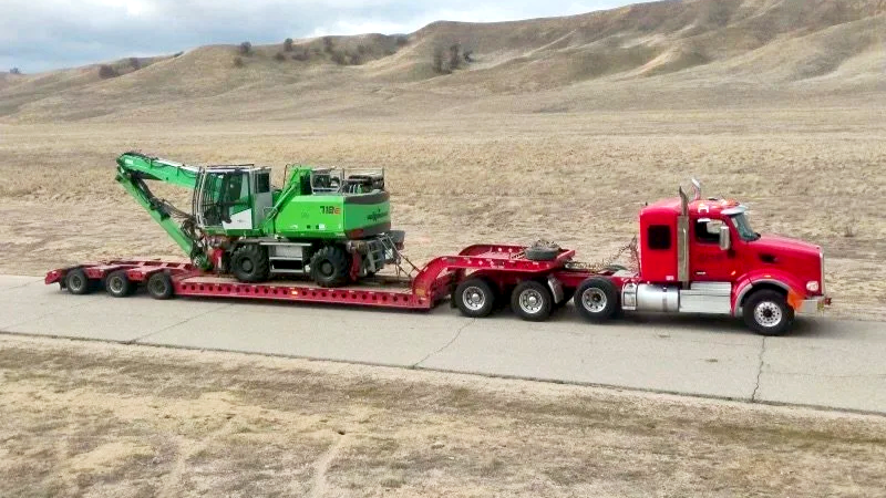 A red semi-truck from H&S Heavy Haul hauling a large green construction excavator on a flatbed trailer on a rural road.