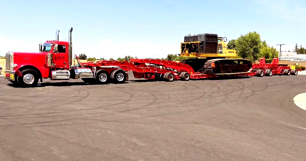 A red semi-truck from H&S Heavy Haul hauling a large black and yellow bulldozer and a red equipment trailer on an asphalt lot with trees in the background.