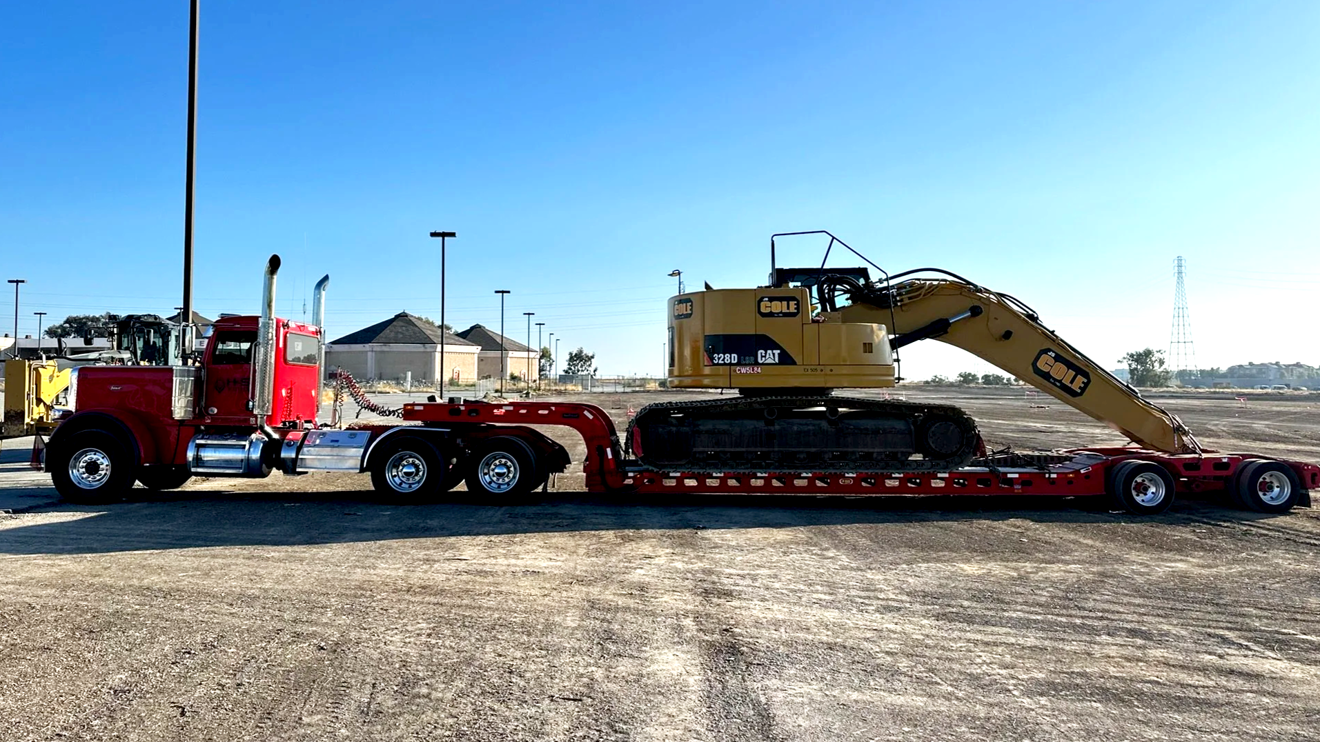 H&S Heavy Haul, A red semi-truck with a yellow excavator loaded on its trailer, parked on a construction site with dirt ground and a few houses in the background.