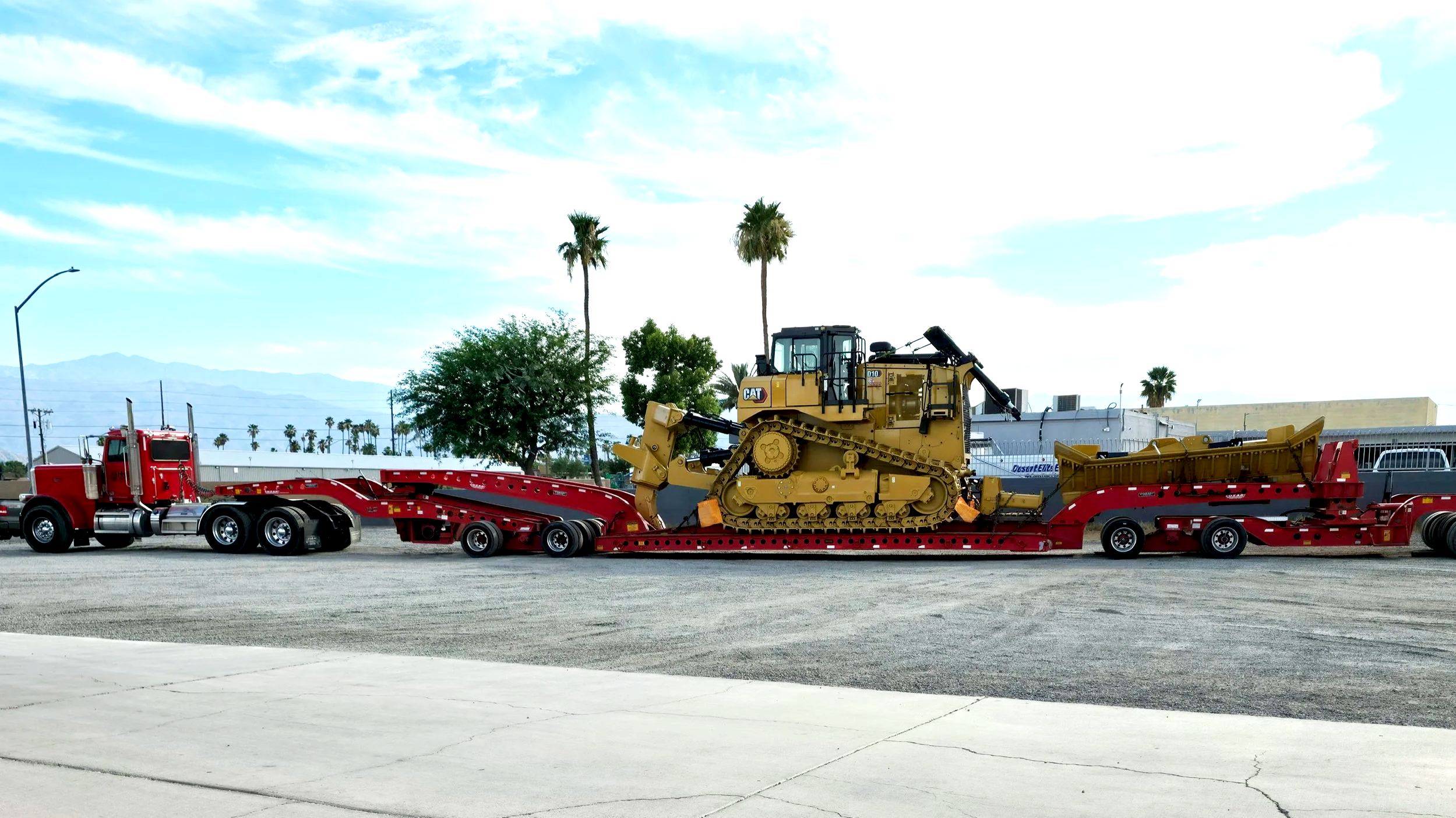 H&S Heavy Haul, Red flatbed tow truck transporting a yellow Caterpillar bulldozer on a road with palm trees and mountains in the background under partly cloudy sky.