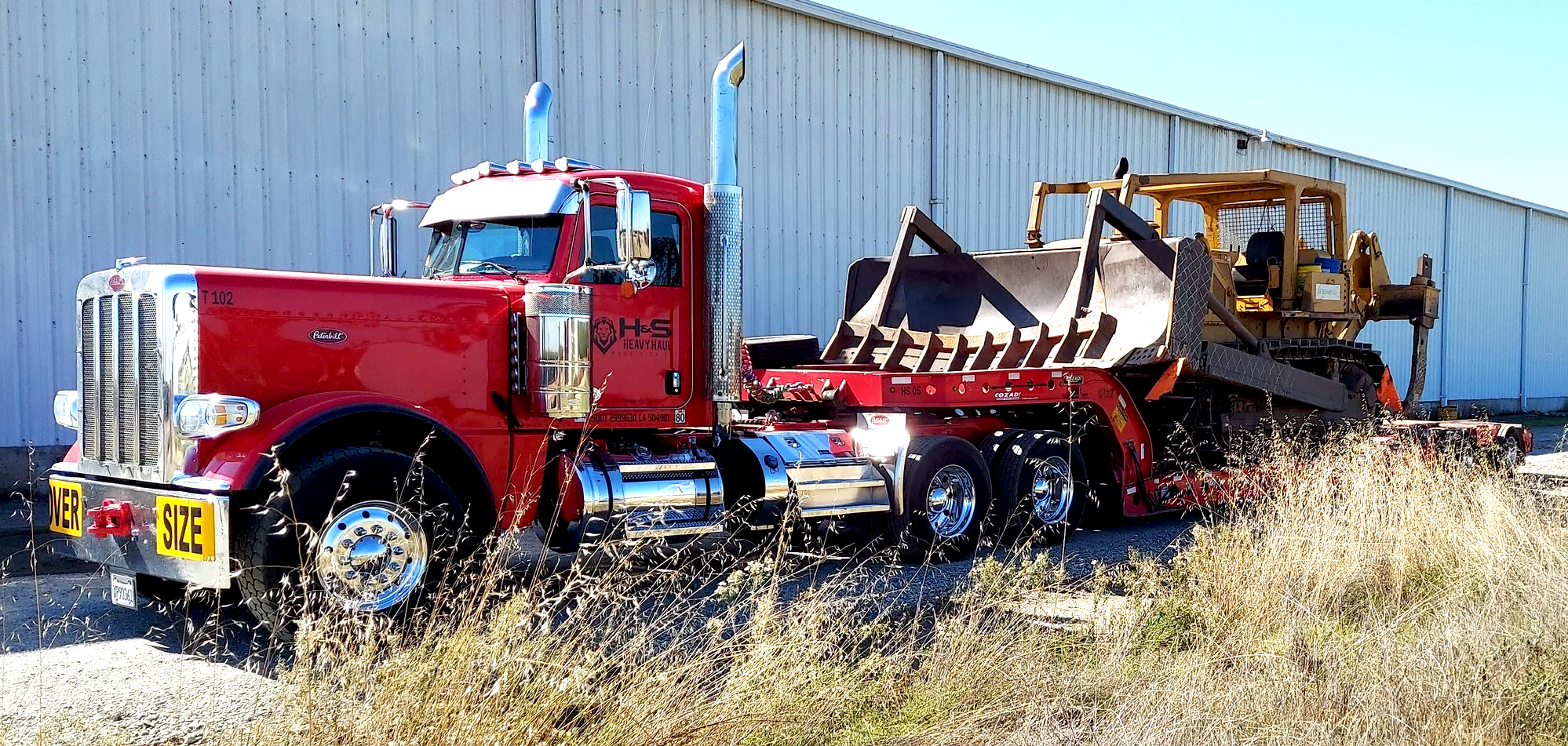 H&S Heavy Haul,  A red Peterbilt heavy-duty truck with a large chrome grille, chrome exhaust stacks, and a flatbed trailer. The truck is hauling a yellow bulldozer with a large front blade, parked beside a light-colored metal building.