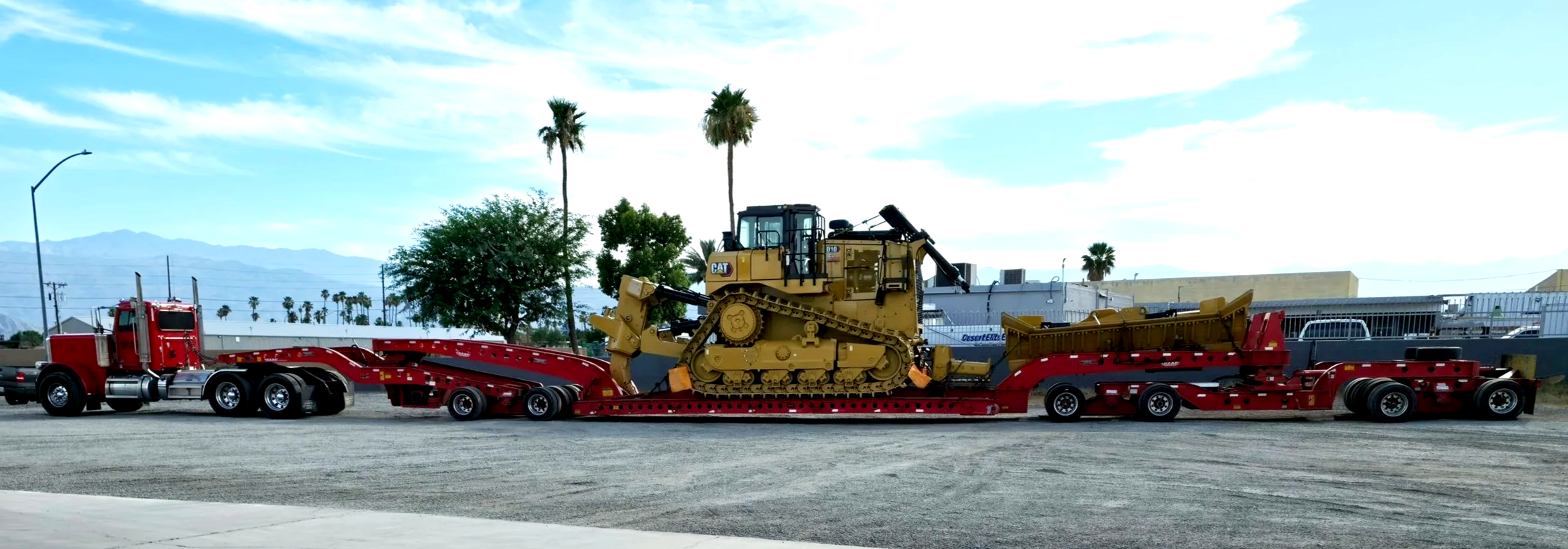 A yellow Caterpillar bulldozer on a red lowboy trailer from H&S Heavy Haul, parked outdoors with palm trees and mountains in the background.