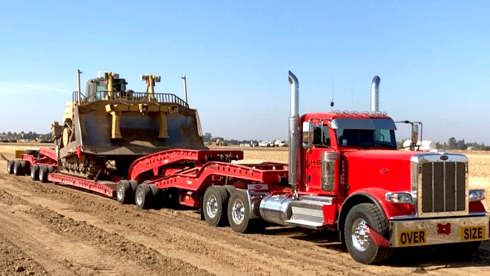 A red heavy-duty truck from H&S Heavy Haul transporting a large bulldozer on a flatbed trailer across a rural dirt road.
