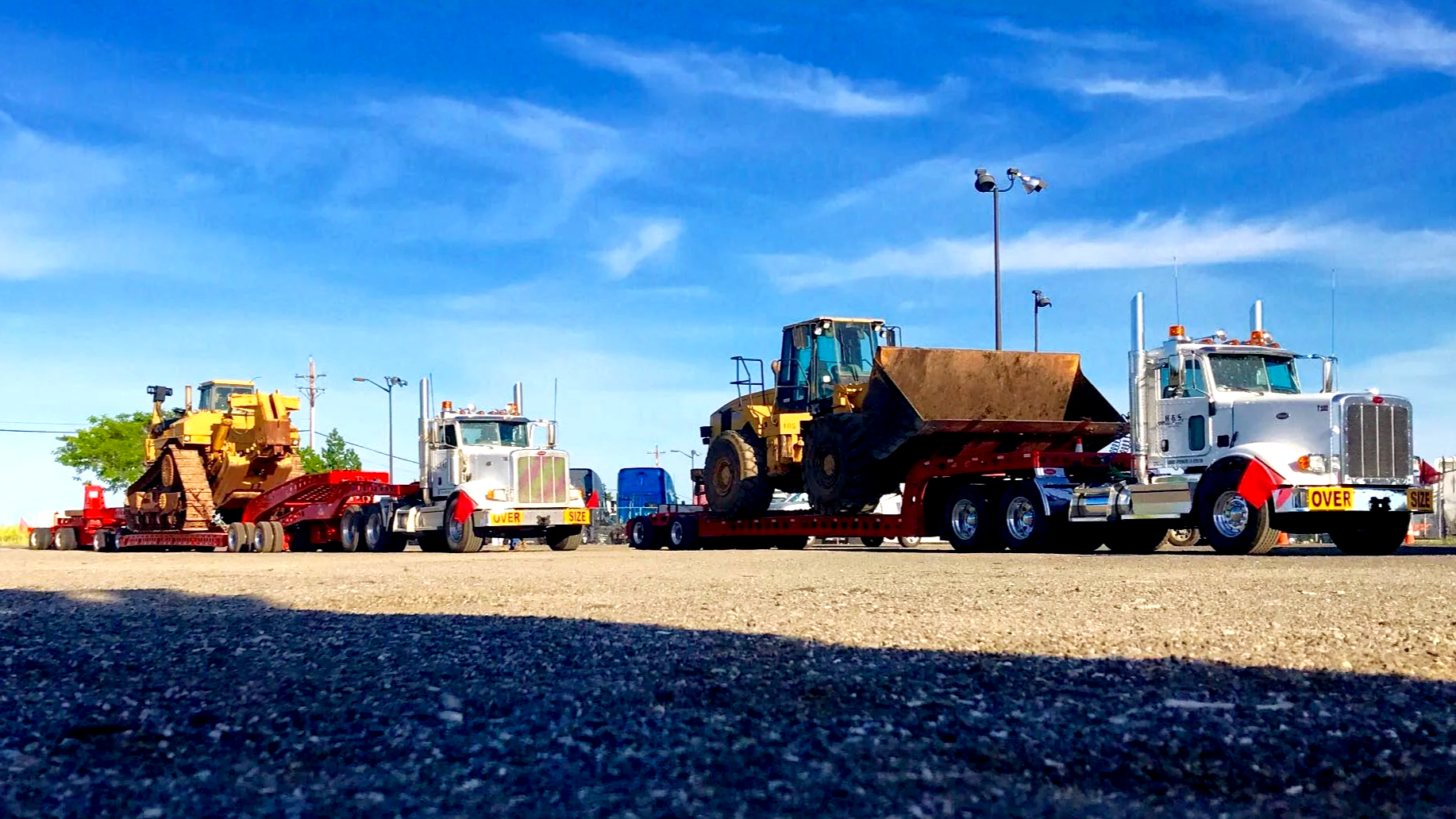 Three trucks from H&S Heavy Haul on a lot carrying construction equipment, under a blue sky with wispy clouds.